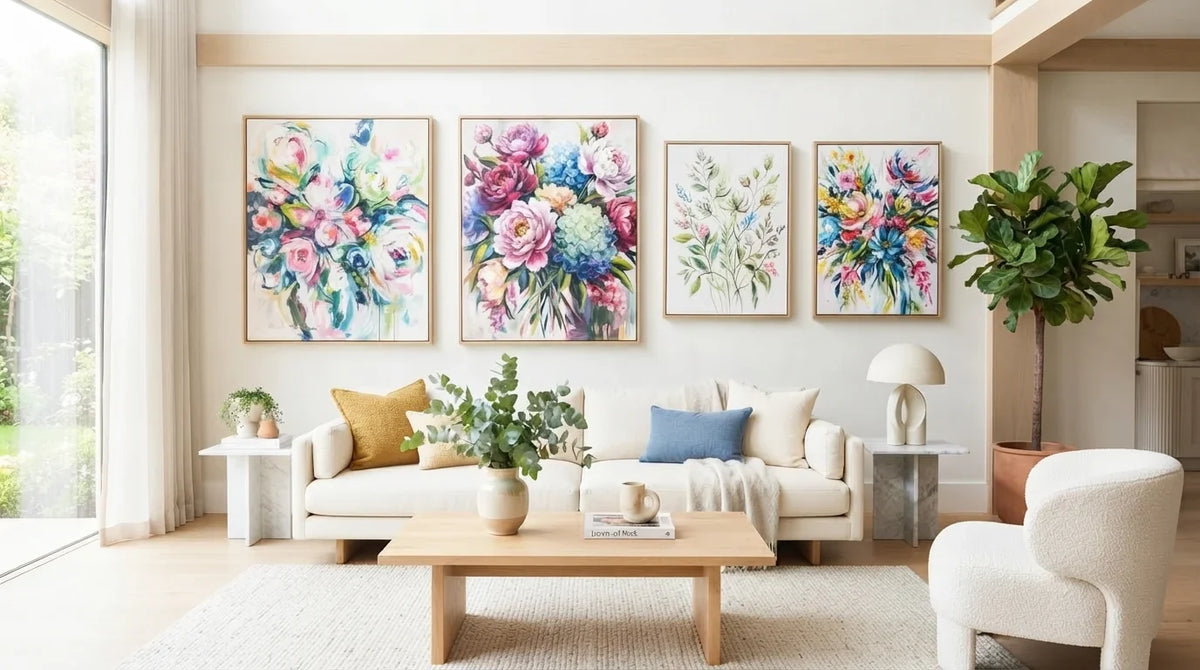 A bright living room with four framed floral art above a cream sofa, a wooden coffee table, and a white armchair beside a tall potted plant.