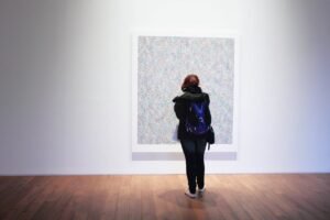 A woman with a backpack observes a colorful painting in an art gallery.